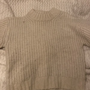 Beige stickad tröja från 157 - Mysig vit-ish stickad tröja från 157 med hög hals och grovstickat mönster. Tröjan har långa ärmar och bred ribbad mudd vid ärmslut och nederkant. Perfekt för kyliga dagar.