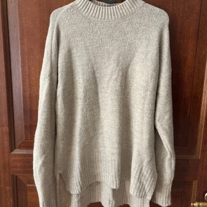 Beige stickad oversized tröja - Mysig beige stickad tröja med ribbad krage, ärmslut och nederkant. Tröjan har en avslappnad oversized passform och är perfekt för kyligare dagar. Enkel och stilren design som passar till det mesta.