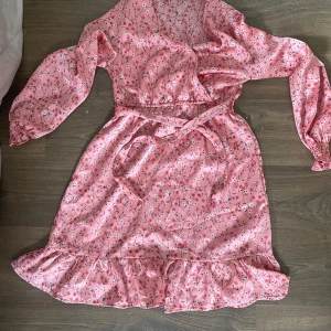 Rosa blommig omlottklänning med volang - Supersöt rosa omlottklänning med blommigt mönster, långa ärmar och volangkant nertill. Klänningen har knytband i midjan och tryckknappar framtill. Perfekt för vår och sommar när du vill känna dig extra fin. Klänningen är i storlek 140 barn. Kan skicka fler bilder 🌸