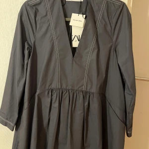 Grå v-ringad klänning från Zara - Snygg grå klänning från Zara med djup v-ringning och markerad midja. Klänningen har långa ärmar med vita kontrastsömmar och ett lätt utställt fall. Perfekt för dig som gillar stilrena och moderna plagg.