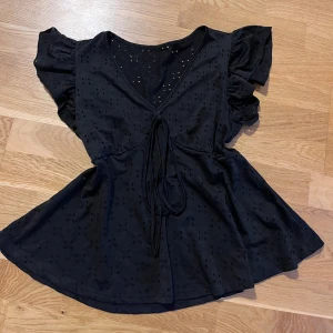 Svart blus med volangärmar och knyt - Säljer en svart blus med v-ringning, volangärmar och knyt framtill. Blusen har broderade hål och en lätt utsvängd passform. Perfekt för dig som vill ha en feminin och trendig topp.