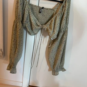 Blommig croppad blus från Pieces - Säljer en ljusbeige croppad blus från Pieces med små blommor i lila och grått. Blusen har långa puffärmar med resår vid ärmslut, knytning framtill och smockad rygg för extra komfort. Perfekt för vår och sommar!