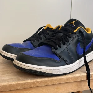 Nike Air Jordan 1 Low svart/blå/gul - Säljer ett par Nike Air Jordan 1 Low med svart läder, blå detaljer och gul insida. Klassisk siluett med svart snörning och vit sula. Väldigt creased men annars bra skick.