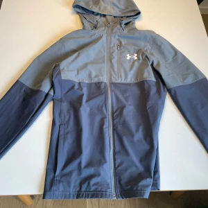Blå vindjacka från Under Armour - Snygg tvåfärgad vindjacka från Under Armour med huva och dragkedja hela vägen. Jackan har en bröstficka med dragkedja och logga på bröstet. Jackan är inprincip helt ny använt 2-3 gånger.