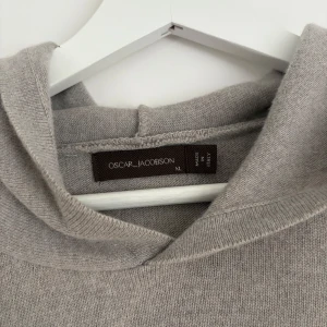 Ljusgrå stickad hoodie från Oscar Jacobson - Mjuk och stilren ljusgrå hoodie från Oscar Jacobson i stickat material. Tröjan har lång ärm, huva och ribbade muddar vid ärmslut och nederkant. Perfekt för en avslappnad och snygg look.