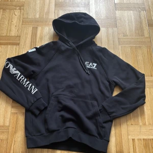 Svart EA7 Emporio Armani hoodie S - Svart hoodie från EA7 Emporio Armani med vit logga på bröstet och stort tryck på ena ärmen. Klassisk känguruficka, huva med snörning och ribbade muddar. Perfekt för en chill och stilren look.