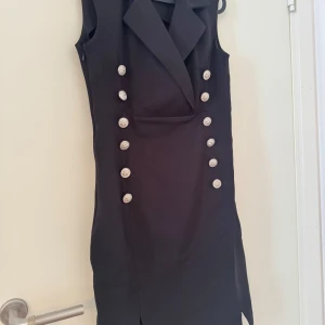 Svart ärmlös kortklänning med knappar - Snygg svart ärmlös kortklänning med markerad krage och dubbla rader av stora silvriga knappar framtill. Klänningen har en figurnära passform och slits nertill, helt ny 