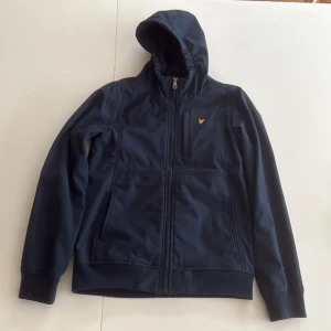 Marinblå vindjacka från Lyle & Scott - Snygg marinblå vindjacka från Lyle & Scott med huva och dragkedja framtill. Jackan har ett litet gult logobroderi på bröstet, praktiska fickor och mjukt fleecefoder på insidan. Perfekt för kyliga dagar och enkel att matcha med det mesta.