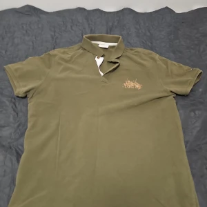 Olivgrön pikétröja från Polo Assn - Säljer en olivgrön pikétröja från Polo Assn med klassisk krage och knappar. Tröjan har korta ärmar, broderad logga på bröstet och en patch på ena ärmen. Perfekt för en stilren och avslappnad look.