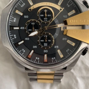 Diesel herrklocka i silver och guld - Stilren herrklocka från Diesel med svart urtavla, flera små urtavlor och detaljer i guld. Armbandet är i rostfritt stål med både silver- och guldfärgade länkar. Klockan har tydliga index och robust design.