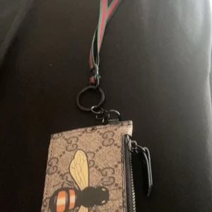 Gucci plånbok med bi-motiv - Säljer en snygg plånbok från Gucci med klassiskt GG-mönster och ett stort bi-tryck på framsidan. Plånboken har dragkedja och grön-röd rem för att hänga runt halsen. Perfekt accessoar för att hålla koll på kort och småsaker.