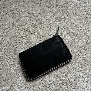 Svart mocka kuvertväska med nitar - Stilren svart kuvertväska i mocka med coola silverfärgade nitar längs kanterna. Väskan har dragkedja upptill och flera fack inuti för att hålla ordning på dina grejer. Perfekt när du vill addera lite edge till din outfit.