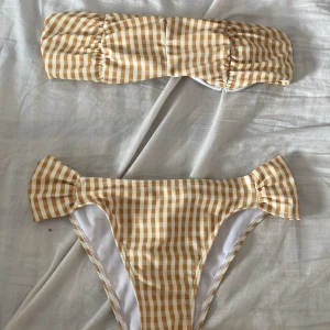 Gul och vit rutig bikini bandeau S - Superfin bikini i bandeaumodell med gul och vit rutig design. Överdelen är axelbandslös och underdelen har söta volangdetaljer på sidorna. Tillverkad i stretchigt material med polyester och spandex. Perfekt för stranden eller poolen i sommar! Helt ny aldrig använt.