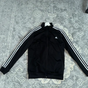 Svart Adidas track jacket S - Svart Adidas track jacket med klassiska vita ränder längs ärmarna och liten Adidas-logga på bröstet. Jackan har hel dragkedja, ståkrage och är tillverkad i polyester. Perfekt för en sportig look eller chill vardagsstil.