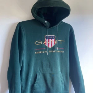 Mörkgrön hoodie från Gant - Snygg mörkgrön hoodie från Gant med broderad logga och sköld på bröstet. Tröjan har huva med snörning och en stor magficka. Perfekt för dig som gillar sportig och klassisk stil. Materialet är mjuk bomullsmix.