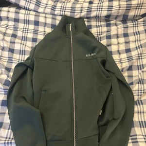 Grön zipjacka från Les Deux - Snygg grön jacka från Les Deux med hel dragkedja framtill och hög krage. Jackan har minimalistisk design, diskret logga på bröstet och två fickor. Perfekt för dig som gillar stilrena plagg med sportig vibe.