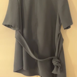 Mörk grön blus med knytband - Stilren mörk grön blus med korta ärmar och ett snyggt knytband i midjan som ger en cool detalj. Perfekt för dig som gillar minimalistisk stil och vill ha något enkelt men ändå trendigt.