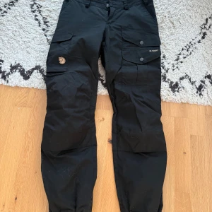 Svarta cargobyxor från Fjällräven - Svarta cargobyxor från Fjällräven med flera praktiska fickor och knappdetaljer. Modell Vidda. Byxorna har normalhög midja och ett kortare ben med justerbara benslut. Passar dig mellan 160-170 cm i längd. Matchande fleecetröja och skaljacka i andra annonser. Storlek 36 = 27-28”