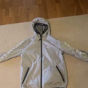 Armani exchange jacka - Snygg ljusgrå vindjacka från Armani Exchange med stor vit AX-logga på ryggen och tryck på ärmarna. Jackan har huva, svarta dragkedjor och detaljer samt två fickor framtill. Perfekt för dig som vill ha en stilren och sportig look.
