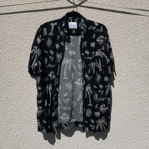 Svart kortärmad skjorta med vit tropisk print från Vaillent - Säljer en svart skjorta från Vaillent med vit tropisk print av palmer, snäckor och drinkar. Skjortan är kortärmad och har knappar framtill. Tillverkad i 100% viskos, och i storlek S.