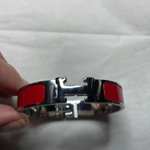 Rött armband från H-rm-s med H-spänne - Säljer ett stilrent armband från H-rm-s i silverfärgad metall med röda emaljdetaljer. Armbandet har det ikoniska H-spännet och är perfekt för att lyfta vilken outfit som helst.