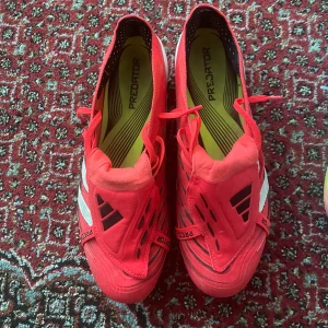 Röda Adidas Predator fotbollsskor - Säljer ett par röda Adidas Predator fotbollsskor med svarta och vita detaljer. Skorna har snörning och är designade för bra grepp och kontroll på planen. Perfekta för dig som vill sticka ut på fotbollsplanen.