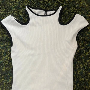 Vit ribbad ärmlös topp från Divided - Säljer en vit ribbad topp från Divided med svarta kanter. Toppen är ärmlös och har unika utskärningar vid axlar och midja för en cool look. Perfekt till sommaren och enkel att styla med jeans eller kjol.