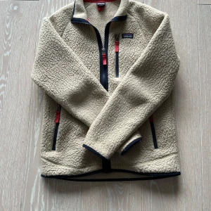 Beige fleecejacka från Patagonia - Säljer en beige fleecejacka från Patagonia med svarta och röda detaljer. Jackan har dragkedja framtill, tre fickor med dragkedja och hög krage. Perfekt för kyliga dagar och outdoor-aktiviteter.