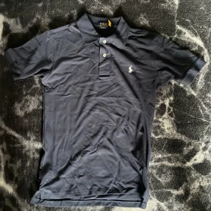 Mörkblå pikétröja från Polo Ralph Lauren - Säljer en mörkblå pikétröja från Polo Ralph Lauren i custom slim fit. Tröjan har korta ärmar, klassisk krage och två knappar framtill. Ikoniska broderade loggan på bröstet. Perfekt för en stilren look.