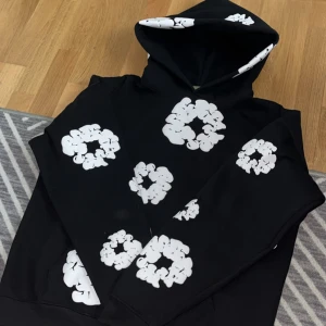 Svart hoodie med vita blommor från Denim Tears - Säljer en svart hoodie från Denim Tears med stora vita blommor tryckta över hela tröjan, även på huvan. Hoodien har en klassisk passform och är perfekt för dig som vill sticka ut med en unik design.