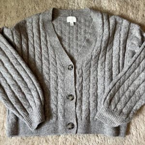 Grå kabelstickad kofta från H&M - Mysig grå kabelstickad kofta från H&M med v-ringning och tre knappar framtill. Koftan har långa ärmar och ribbade muddar vid ärmslut och nederkant. Perfekt för lager-på-lager och enkel att matcha med olika outfits.