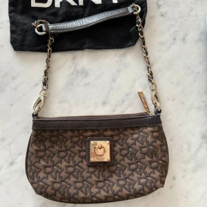 Brun axelväska från DKNY med kedjerem - Säljer en brun axelväska från DKNY med kedjerem och läderdetaljer. Väskan har ett monogrammönster med DKNY-logga över hela ytan, dragkedja upptill och en guldfärgad platta framtill. Perfekt för dig som gillar stilrena accessoarer med lyxig känsla.