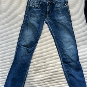 Replay Ambass blå jeans herr 30/32 - Snygga Replay Anbass jeans i klassisk blå tvätt med lätt slitning. Modellen har smal passform och raka ben, fem fickor och detaljerade sömmar. Perfekta för dig som gillar stilrena och moderna jeans med en bekväm känsla.