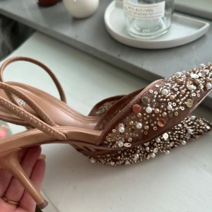 Bruna pumps med pärlor och paljetter - Säljer ett par bruna pumps med spetsig tå och öppen häl. Skorna är dekorerade med pärlor och paljetter i olika storlekar för en riktigt glittrig look. Klackhöjd är medel och materialet känns syntetiskt. Använda en gång 