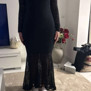 Svart spetsklänning med öppen rygg - Elegant svart klänning i spets med lång ärm och en stilfull öppen rygg. Klänningen har en tight passform och når ner till vaderna med en transparent spetsdetalj vid fållen.