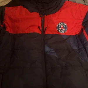 Röd och marinblå jacka från Paris Saint-Germain - Säljer en snygg röd och marinblå jacka från Paris Saint-Germain. Jackan har en dragkedja framtill och klubbens logga på bröstet. Perfekt för fotbollsfans som vill visa sitt stöd med stil.