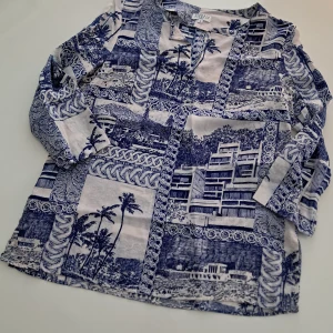 Blå och vit mönstrad blus från Claudie Pierlot - Säljer en snygg blus från Claudie Pierlot i SIDEN av högsta kvalitet, med ett unikt blått och vitt mönster som föreställer palmer och byggnader. Blusen har långa ärmar och en avslappnad passform, perfekt för en stilren look. Liten I storleken, därför 34 (XS).