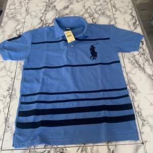 Blå randig pikétröja från Ralph Lauren - Säljer en stilren blå pikétröja från Ralph Lauren med svarta ränder och det klassiska logotypbroderiet på bröstet. Tröjan har en krage och knappar framtill. Perfekt för en avslappnad men ändå snygg look.