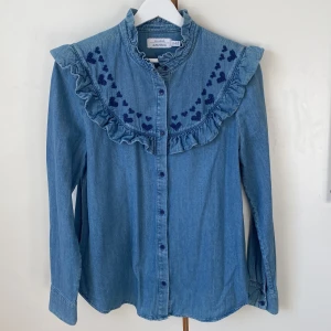 Blå denimblus från & Other Stories - Supersöt blå denimblus från & Other Stories med volangdetaljer och broderade hjärtan i korsstygn på framsidan. Skjortan har långa ärmar och knappar framtill. Längd 70. Storlek 40. 