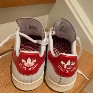 Adidas Stan Smith sneakers med röd detalj - Snygga Adidas Stan Smith sneakers i vitt med röda detaljer. Skorna har en klassisk design med snörning och en unik innersula med hjärtmönster. Perfekta för en stilren look. Skicket är väldigt bra, används 1 gång
