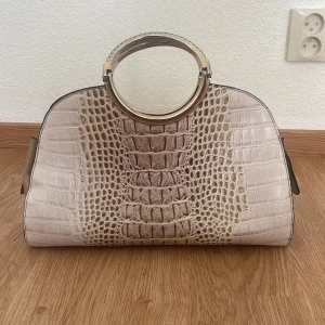 Beige handväska med krokodilmönster - Elegant beige handväska med krokodilmönster och rundade handtag i metall. Väskan har en dragkedja framtill och flera fack inuti för praktisk förvaring. Perfekt för att ge en lyxig touch till din outfit. 
