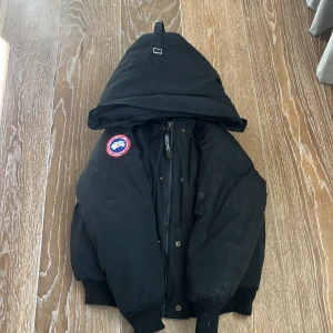 Svart dunjacka från Canada Goose - Säljer en svart dunjacka från Canada Goose med huva och dragkedja. Jackan har en broderad logotyp på ärmen och knappar framtill. Perfekt för kalla vinterdagar. Saknar päls runt luvan