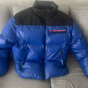 Blå och svart dunjacka från Prada - Säljer en snygg blå och svart dunjacka från Pradas kollektion linea rossa med dragkedja och hög krage. Jackan har en pufferdesign och är tillverkad i polyamid med avancerad vadderingsteknik. Perfekt för kyliga dagar! Nypris 34900kr och köpt på Prada Stockholm Birger jarlsgatan. Jackan har även en inbyggd luva i kragen. 