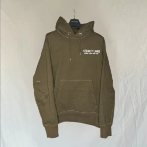 Olivgrön hoodie från Helmut Lang - Säljer en snygg olivgrön hoodie från Helmut Lang med tryck på bröstet och ryggen. Tröjan har en klassisk känguruficka och justerbar huva med snören. Perfekt för en avslappnad stil. Utan cargo banden på armarna nypris runt 3