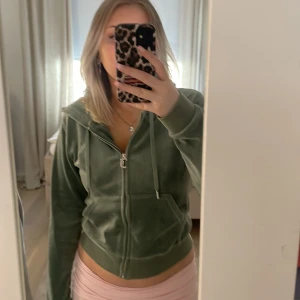 Grön velourjacka från Juicy Couture - Säljer en snygg grön velourjacka från Juicy Couture. Jackan har dragkedja och huva, perfekt för en avslappnad stil. Den är långärmad och har en mjuk känsla som gör den bekväm att bära.