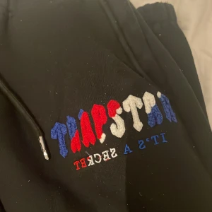 Trapstar tracksuit får med tröjan och byxorna  - Svarta mjukisbyxor med broderad text i rött, vitt och blått. Byxorna har en elastisk midja med snörning och dragkedjor vid bensluten för justerbar passform.