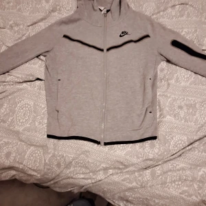 Grå huvtröja från Nike - Säljer en grå huvtröja från Nike med dragkedja och svarta detaljer. Tröjan har en huva och långa ärmar, perfekt för en sportig look. Den är tillverkad i ett mjukt material för extra komfort.