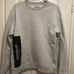 Grå sweatshirt från Dondup - Säljer en stilren grå sweatshirt från Dondup med svart tryck på sidan. Tröjan har långa ärmar och en rund halsringning. Perfekt för en avslappnad look.