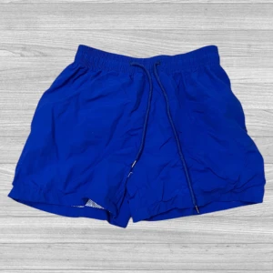 Blå badshorts med snörning - Snygga blå badshorts med elastisk midja och snörning för perfekt passform. Shortsen har en praktisk bakficka med knapp och är tillverkade i ett lätt och snabbtorkande material.