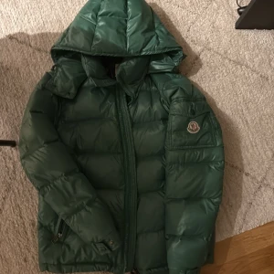 Grön dunjacka från Moncler - Säljer en snygg grön dunjacka från Moncler med huva och dragkedja. Jackan har en quiltad design och Moncler-logga på ärmen. Perfekt för kyliga dagar!